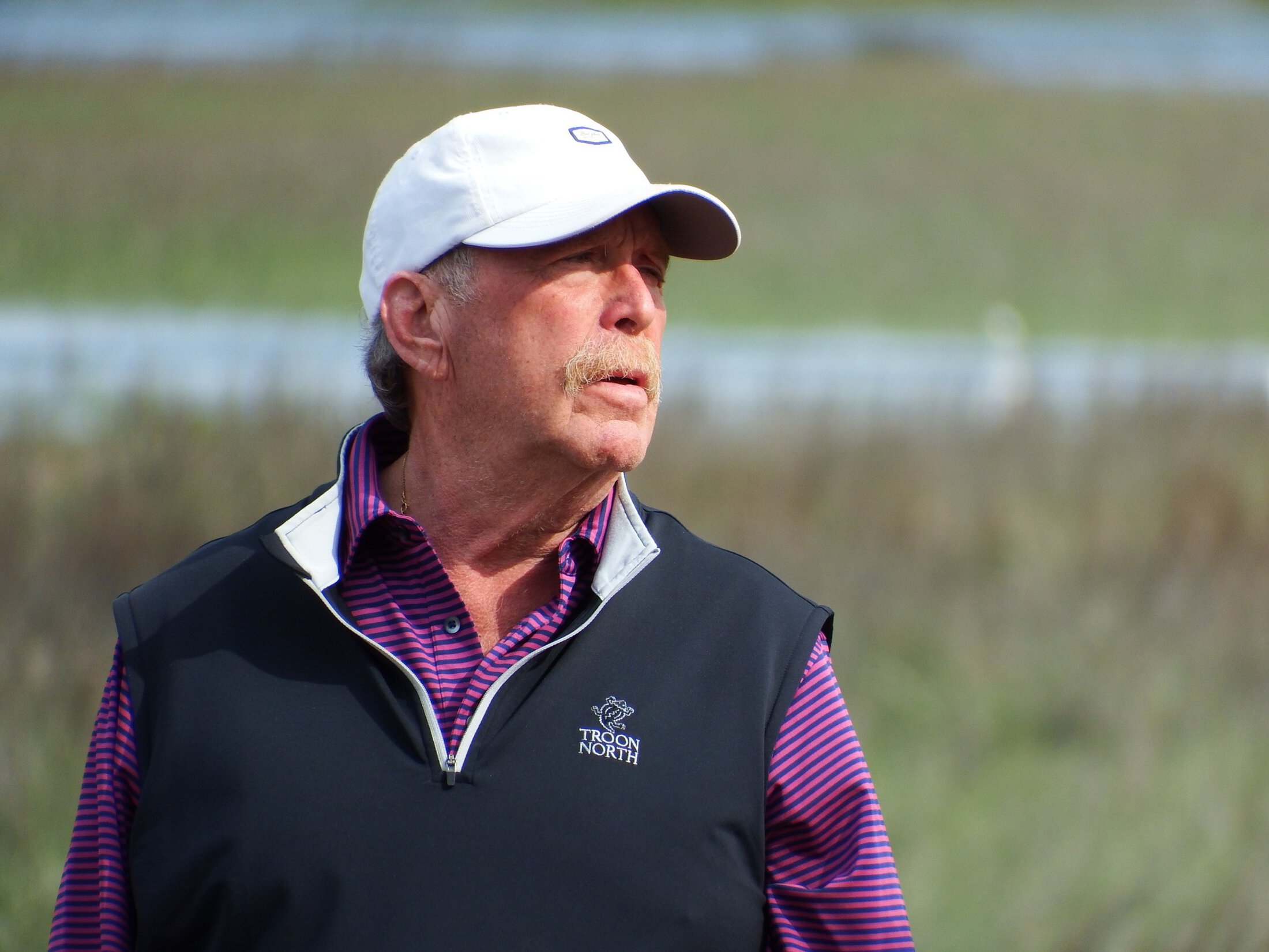 A man with a moustache stands outside. He is wearing a white ballcap, a blue and purple striped golf shirt and a vest that says Troon North.