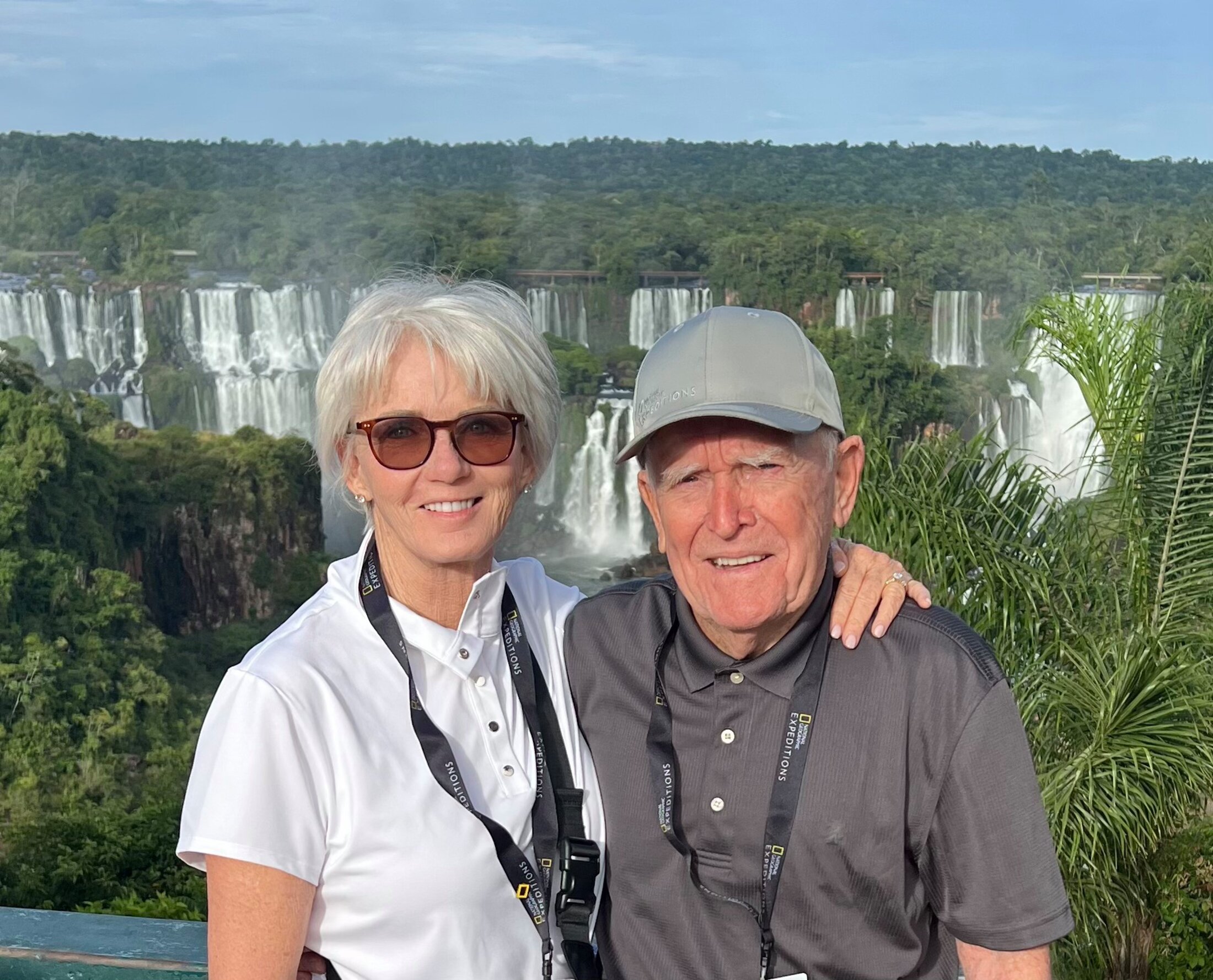 Two older adults smile at the camera with a dramatic waterfall and lush rain forest in the background.