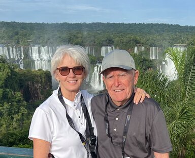 Two older adults smile at the camera with a dramatic waterfall and lush rain forest in the background.
