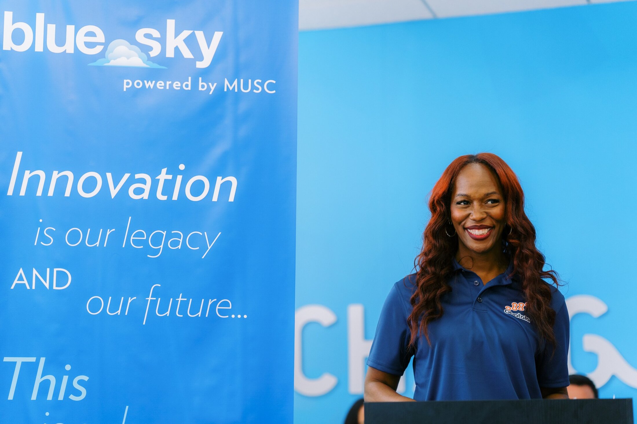A woman with long, reddish hair wearing a blue polo shift smiles as she stands behind a podium. In the back ground are the words 
