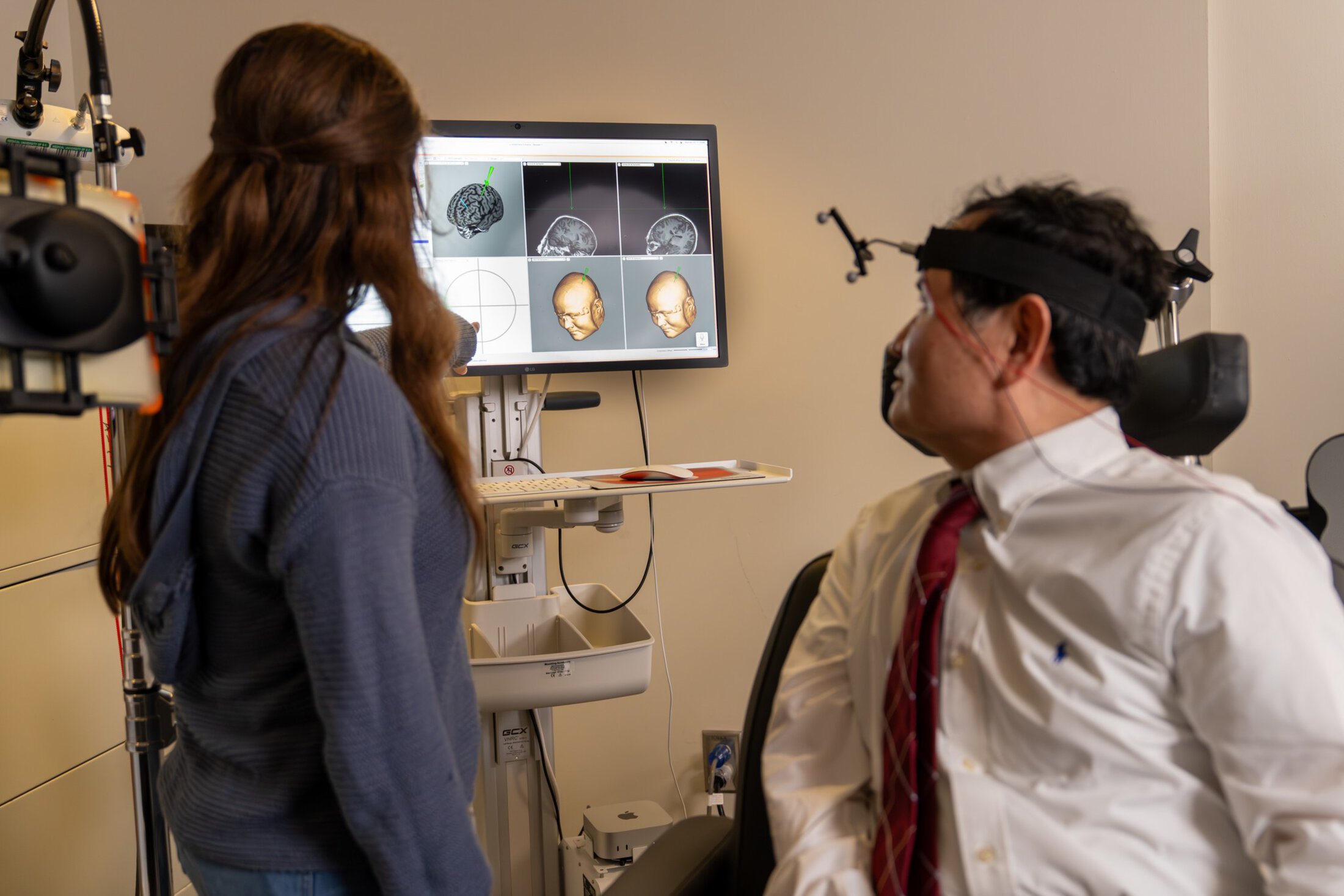 A seated man wearing a strap with wires around his forehead twists in his chair to look at a computer monitor showing brain images.