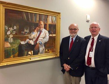 Two men in suits stand next to a portrait that looks like a painting of the man on the left. The person in the portrait is wearing a white doctor's coat, seated in a chair by a table with a coffee cup, eyeglasses and flowers.