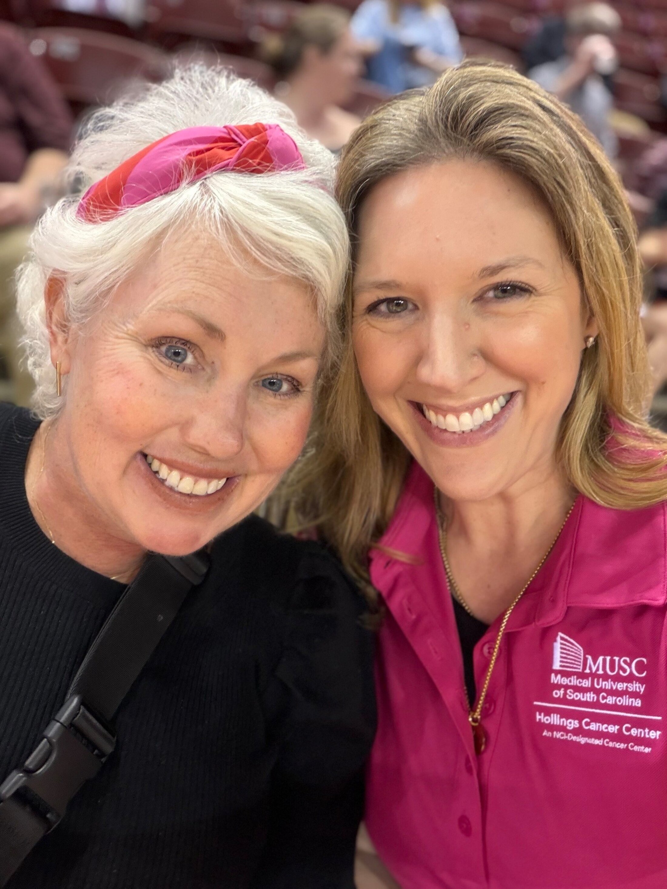 Two women, one wearing a pink headband and the other in a pink shirts that says Hollings Cancer Center, lean together to pose for a photo.