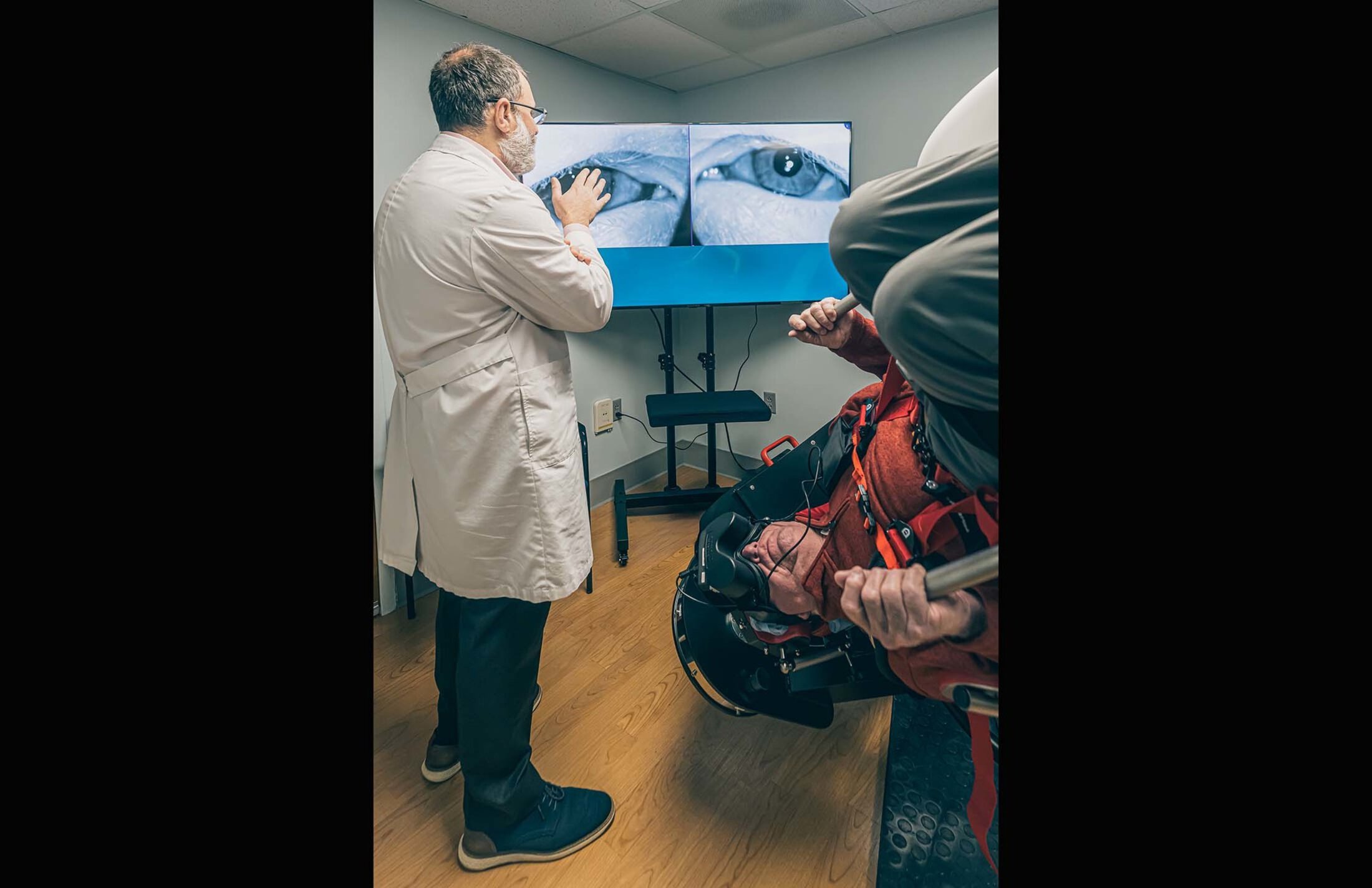 Man in white doctor's coat stands on the left side gesturing to a screen showing two eyes. On the right, a man is nearly upside down in a chair that he's strapped into.