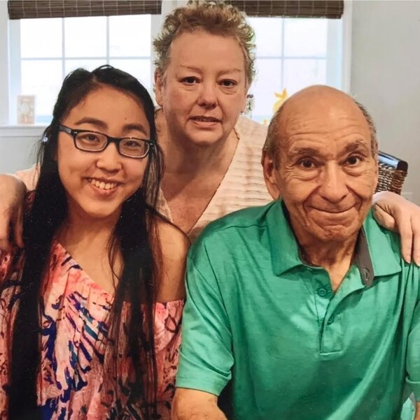 Three people posing together indoors in front of a window. A young woman with glasses sits beside an older man in a green polo shirt, while a woman stands behind them with her arms around their shoulders. All three are smiling at the camera.