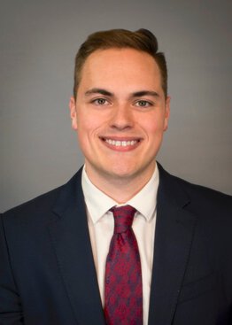 Matthew Triano wearing a dark blue sport coat and dress shirt and red tie