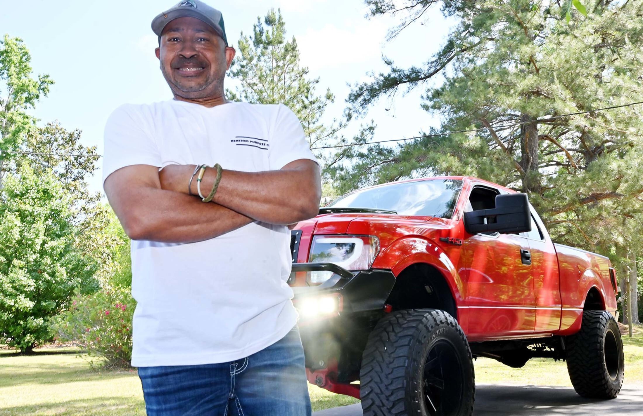 A smiling man wearing a ballcap, white t-shirt and jeans crosses his arms over his chest. There is a red truck behind him.