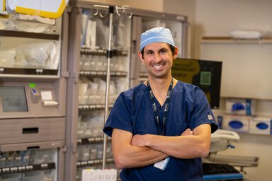 Smiling male healthcare professional in surgical scrubs and cap standing with arms crossed in a clinical or operating room setting, with medical equipment and supplies in the background.