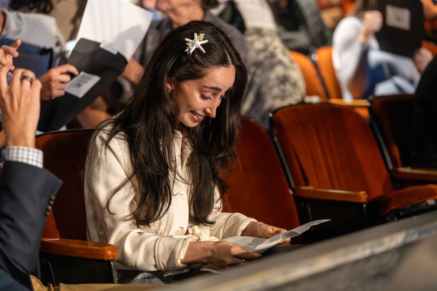 A young woman sitting in theater seating smiles as she reads a letter.