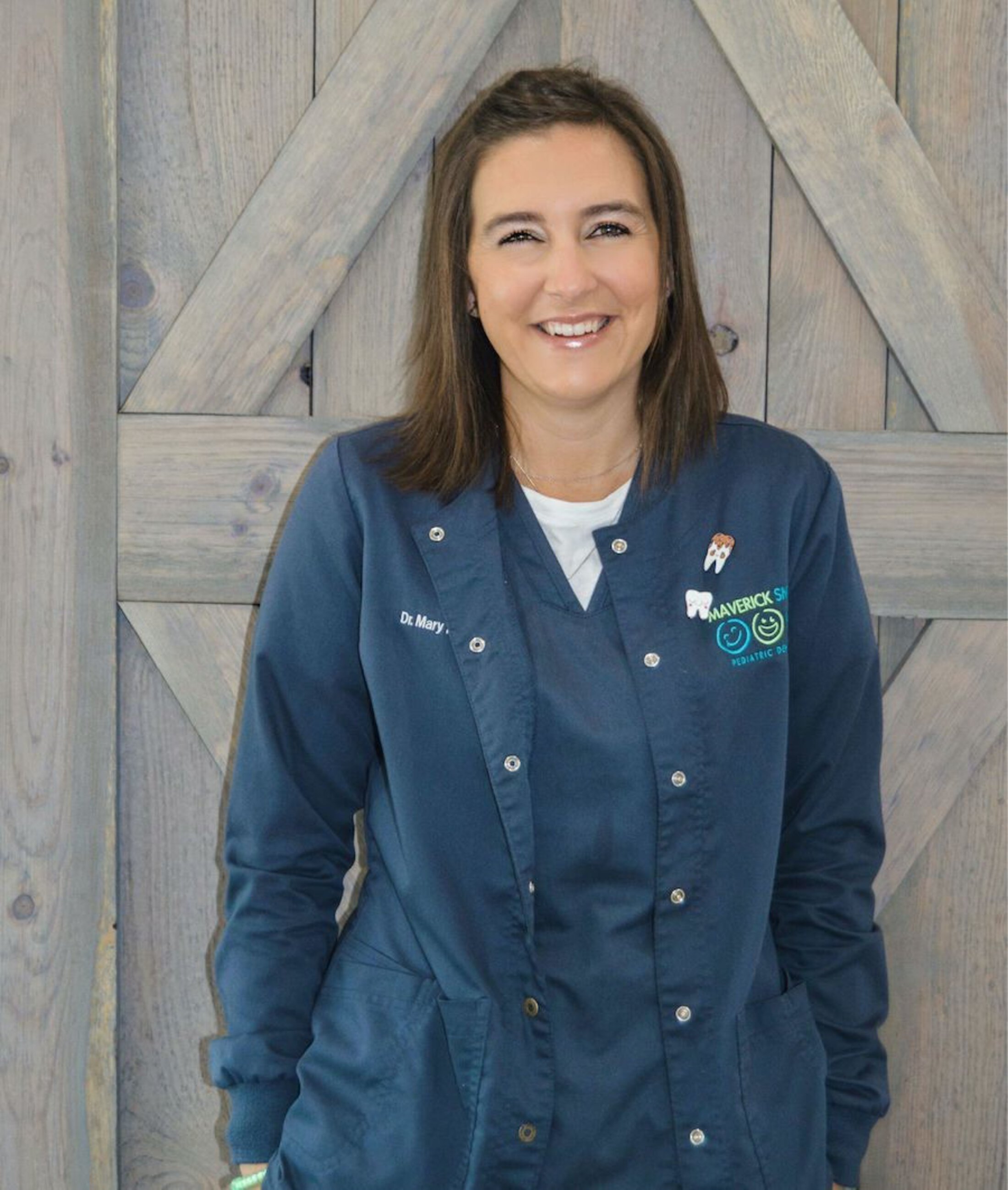 Portrait of a smiling woman wearing blue dental scrubs, standing in front of a rustic wood-paneled wall.