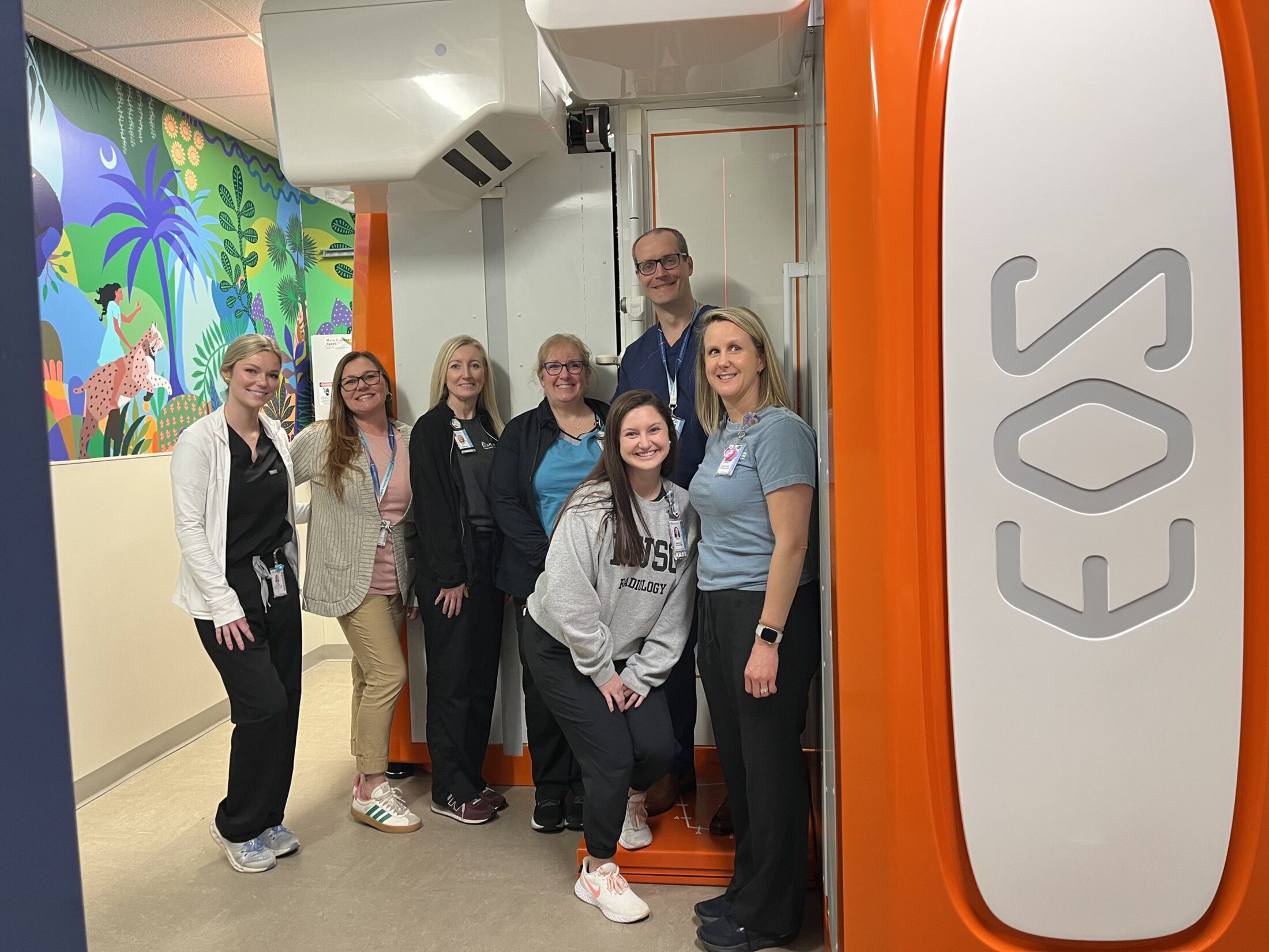 A group of seven healthcare staff members — wearing scrubs, lab coats, and casual clinical attire — smile together beside a large orange EOS imaging machine. A colorful jungle mural decorates the wall behind them.