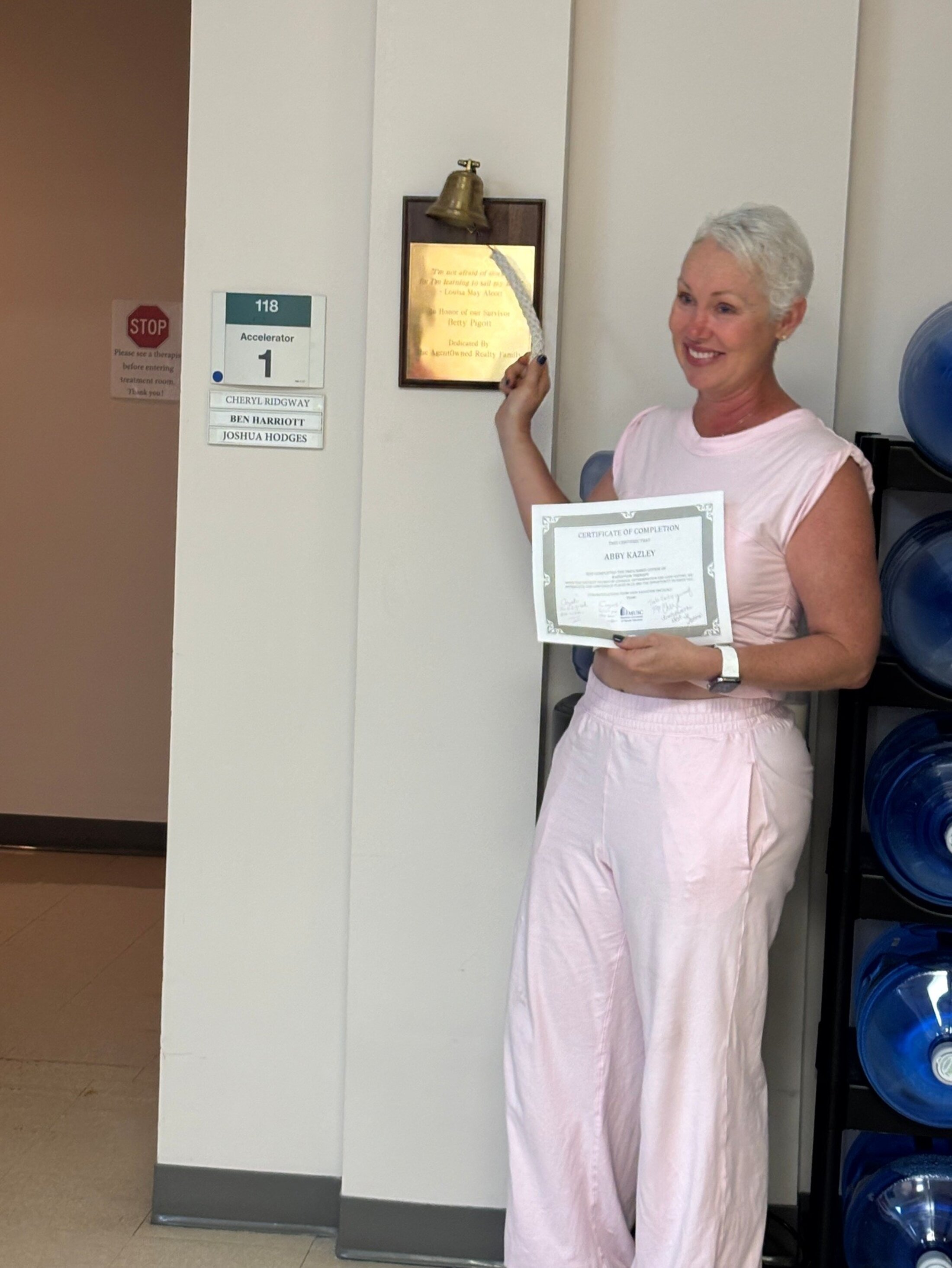 A woman in pale pink blouse and pants holds a certificate and rings a bell.