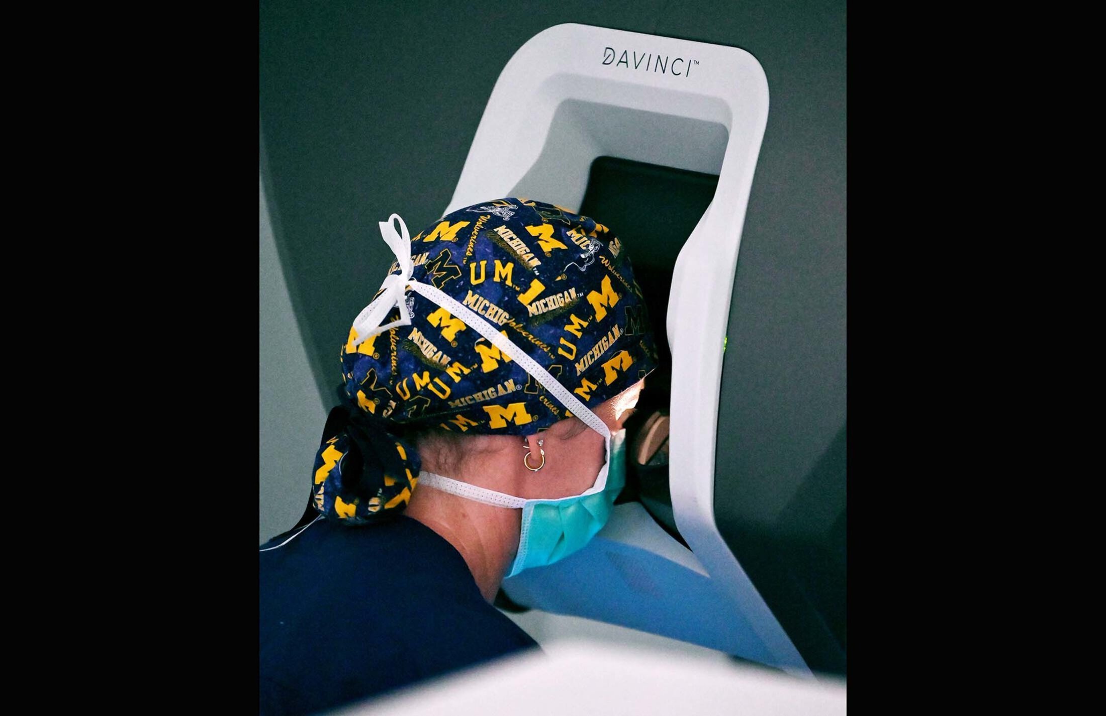 Side of a woman's head. She's wearing a hair covering with University of Michigan logos. She has a face mask and is leaning into a white opening that says DaVinci on the top of it.