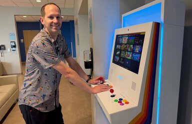 Man with hands on video game machine. He's smiling and facing the camera. He is in what looks like a waiting room. Theman is wearing a shirt with people all over it and black pants.