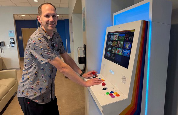 Man with hands on video game machine. He's smiling and facing the camera. He is in what looks like a waiting room. Theman is wearing a shirt with people all over it and black pants.