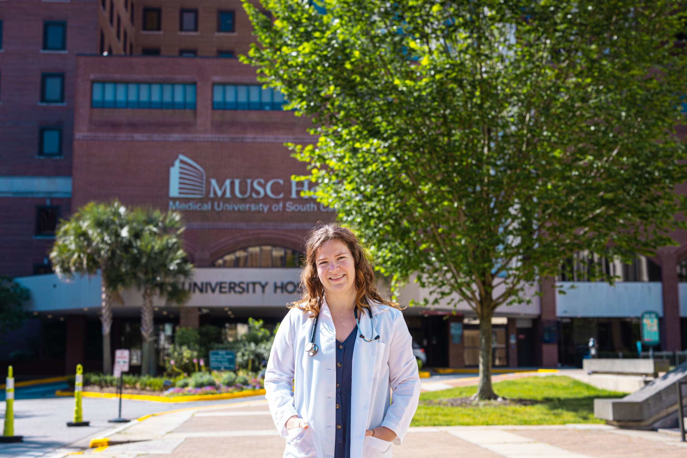 Rachel Burge, a medical student and recent Ph.D. graduate, stands outside MUSC University Hospital wearing a white coat and stethoscope, smiling toward the camera on a sunny day. Trees and the Medical University of South Carolina campus are visible in the background.