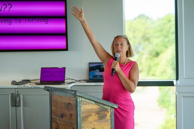 A breast cancer survivor in a bright pink dress speaks confidently at a podium.