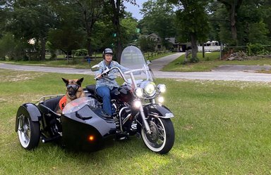 A woman wearing a helmet and a dog wearing goggles sit on a black Harley motorcycle with a sidecar.