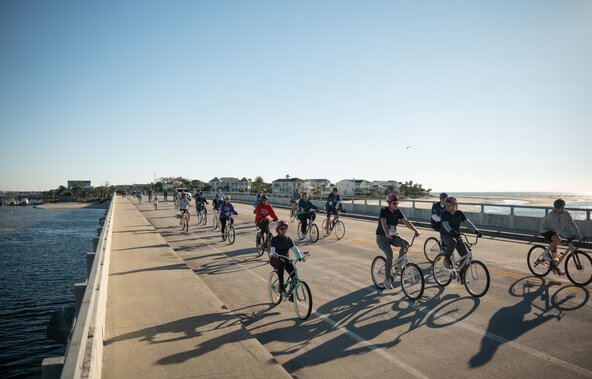 a large group of cyclists ride over a bridge with beach houses behind them