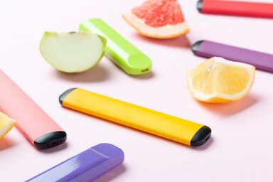 Brightly colored vape pens next to slices of equivalent fruit pop on a pastel pink background.