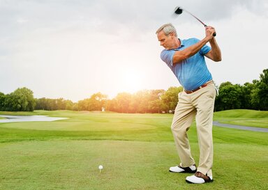 A man with gray hair wearing a blue polo shirt, khakis and white golf shoes prepares to hit a golf ball.