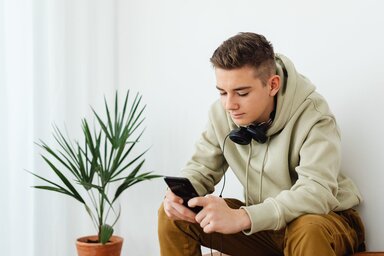 Young man with short hair leans forward to look at his smartphone. there are headphones around his neck, he is wearing a hoodie and brown pants. A small green plant sits beside him.