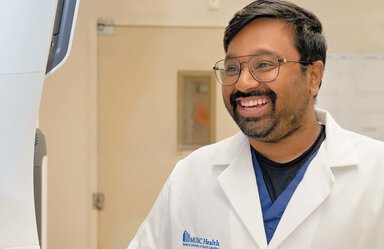 Closeup of a smiling man wearing eye glasses and a white doctor's coat. He has dark hair and a beard.
