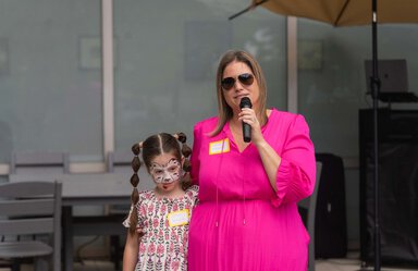 A girl with long brown hair is wearing a dress and face paint. She is being held by a woman wearing a bright pink dress aviator sunglasses. The woman is speaking into a microphone.