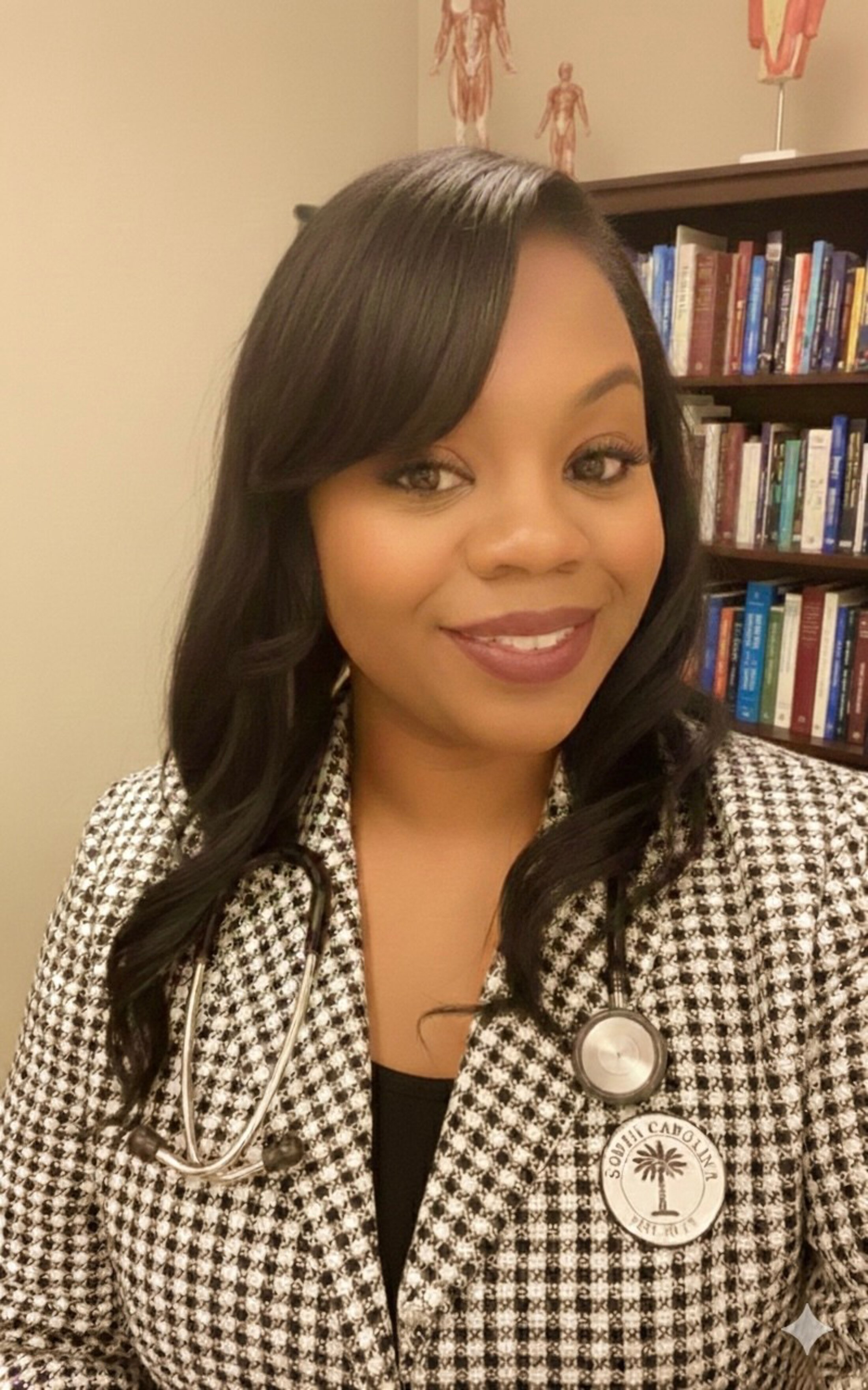 Woman with long dark hair smiling at the camera in an office setting, wearing a patterned jacket and a stethoscope around her neck, with bookshelves visible behind her.