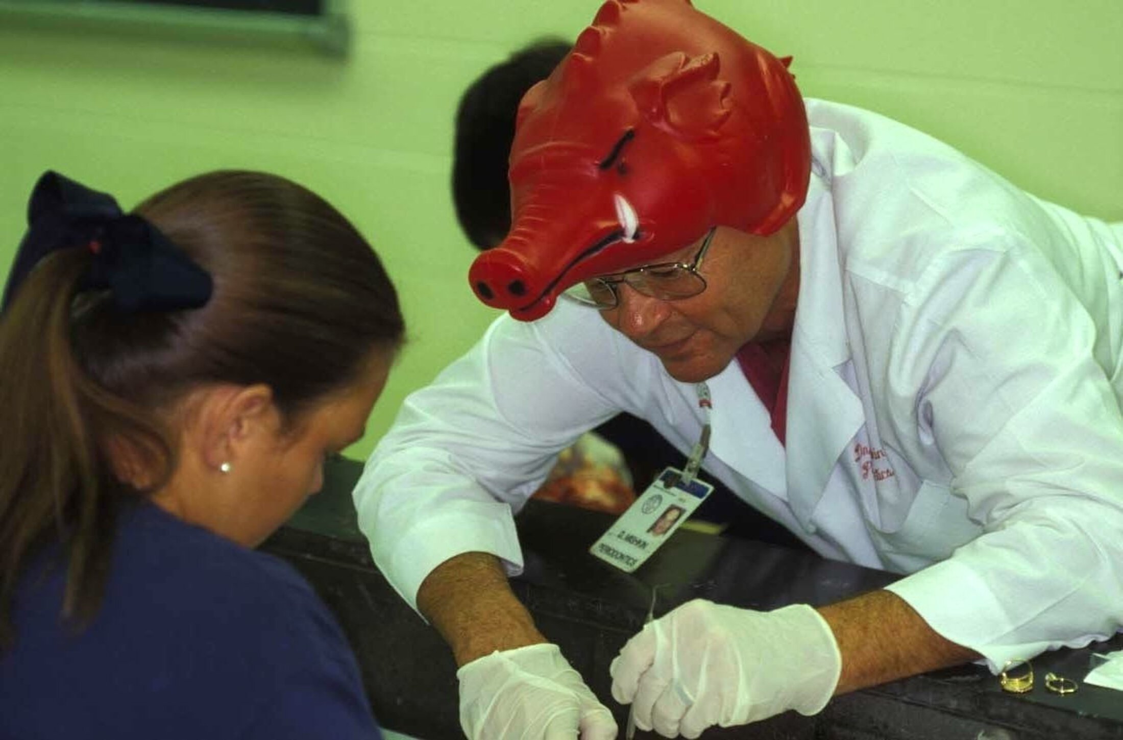Dr. Mishkin leans over a lab station to inspect a students work. He is wearing a hat in the shape of a pig mascot.
