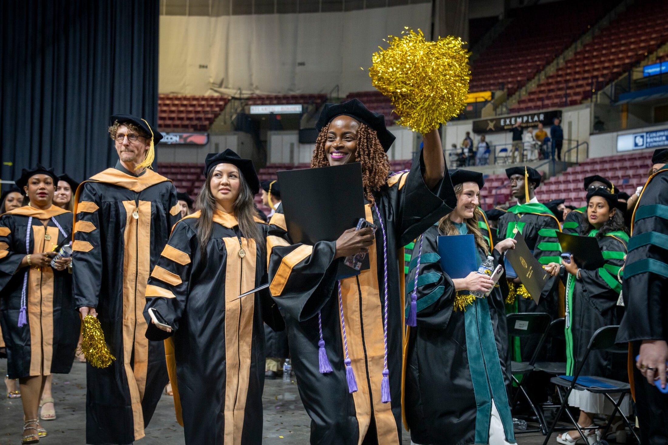 Graduates in academic regalia process through an indoor arena during a commencement ceremony. In the center, a smiling graduate in a black doctoral gown with gold trim raises a gold pom-pom while carrying a diploma cover. Other graduates in doctoral robes walk alongside and behind her, some holding programs and water bottles, with stadium seating visible in the background.