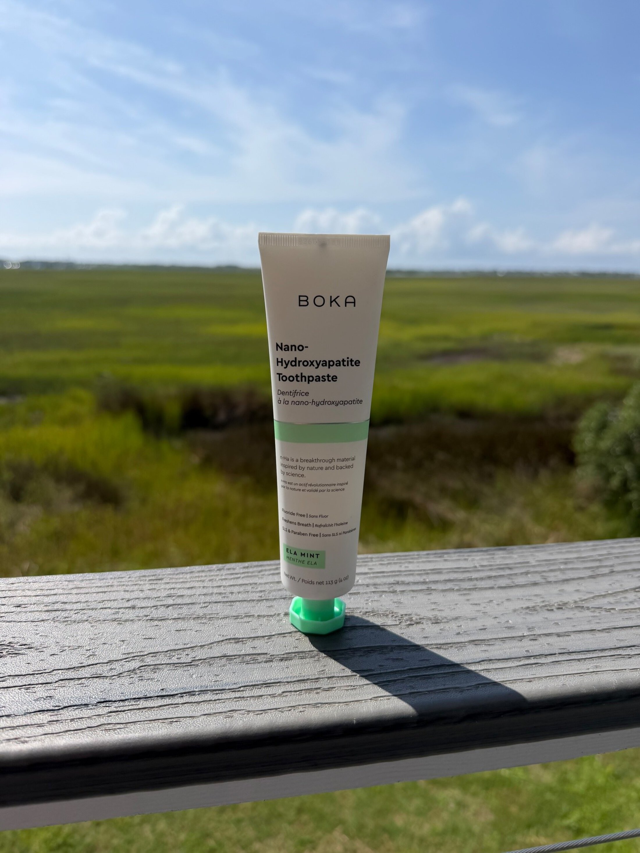 A white tube of toothpaste sits on a rail outside with the marsh and blue sky as a bakdrop. The tube says BOKA Nano-hydroxyapatite toothpaste. ELA mint.
