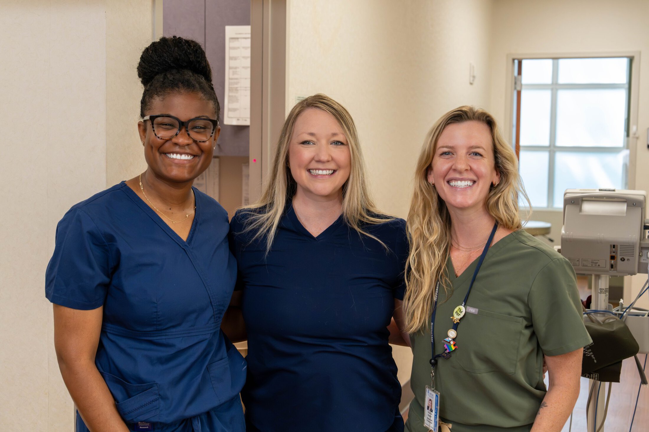 Three women in scrubs pose in a clinic hallway.
