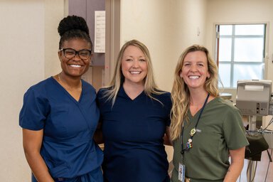 Three women in scrubs pose in a clinic hallway.