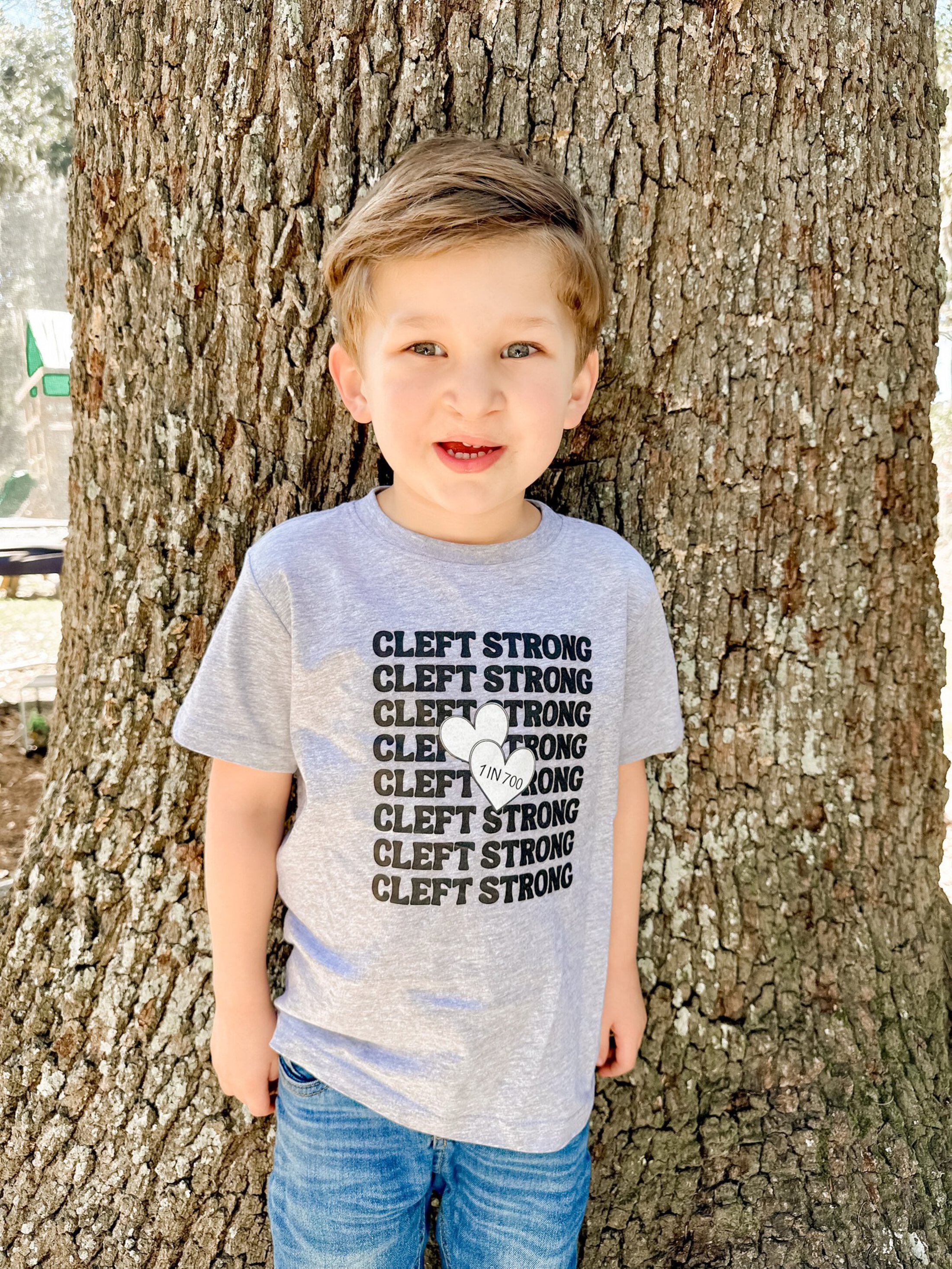 Photograph of a young child standing in front of a large tree trunk outdoors. The child wears a gray t-shirt with repeated black text 