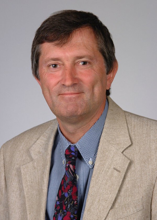 Headshot of a man wearing a tan jacket, blue and white checked shirt and colorful necktie.