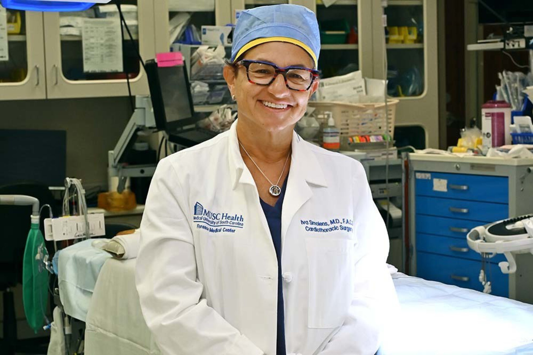 Dr. Smolens, smiling wearing a lab coat inside a hospital room with a hospital bed