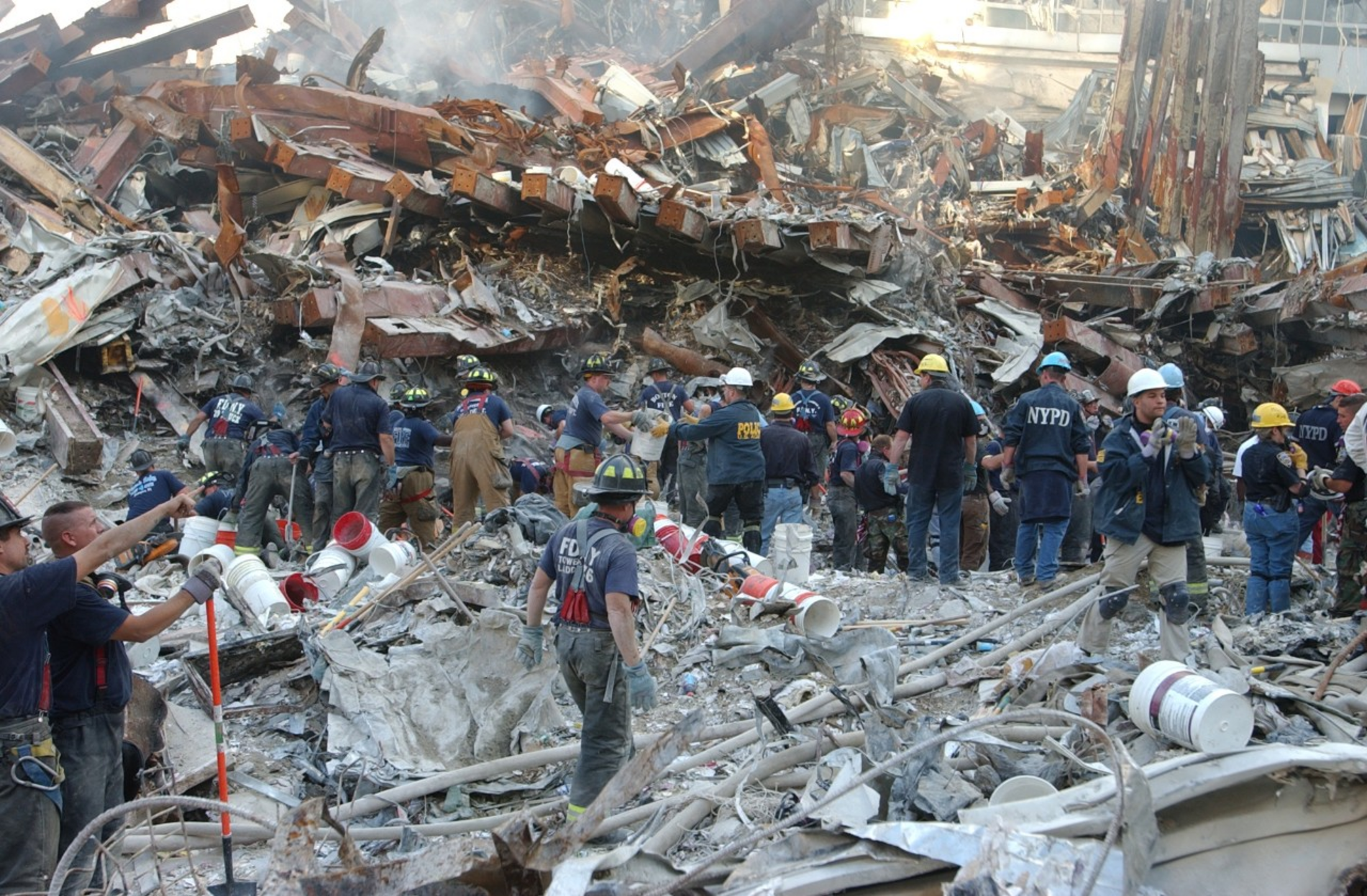 A huge pile of building debris. There is smoke or ash in the air. A large number of people in NYPD and FDNY shirts and jackets are working with it or moving toward it.
