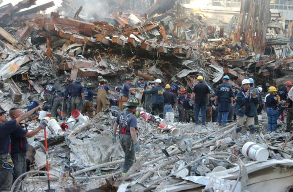 A huge pile of building debris. There is smoke or ash in the air. A large number of people in NYPD and FDNY shirts and jackets are working with it or moving toward it.