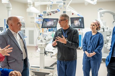 A man wearing glasses and hospital scrubs gestures while talking to a man wearing a suit. A woman in scrubs is behind the speaker, listening.