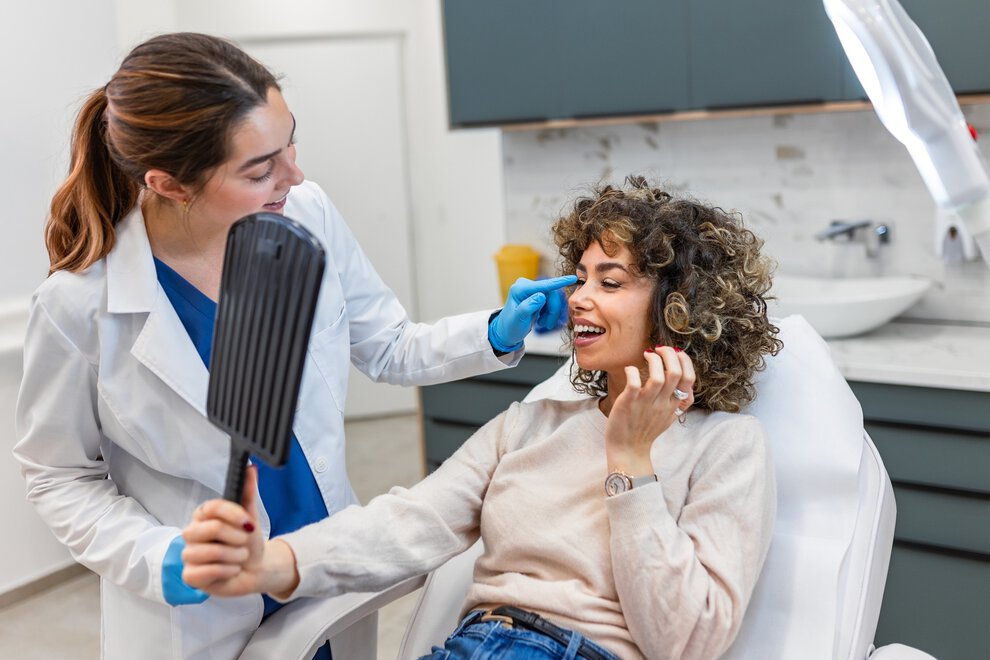 Happy woman holding a mirror and looking at the results after a successful botox treatment. Aesthetic medicine concept. Female surgeon talking to a smiling patient in a professional medical clinic.