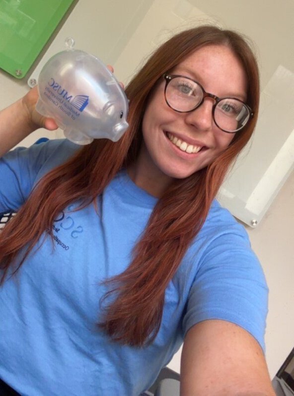 Smiling student wearing glasses holds a clear MUSC-branded piggy bank during a financial literacy outreach event.