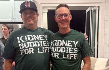 Two men smile as they stand together in a kitchen. The one on the left is wearing a ball cap. The one on the right is wearing round eyeglasses. Both are wearing t-shirts that say 