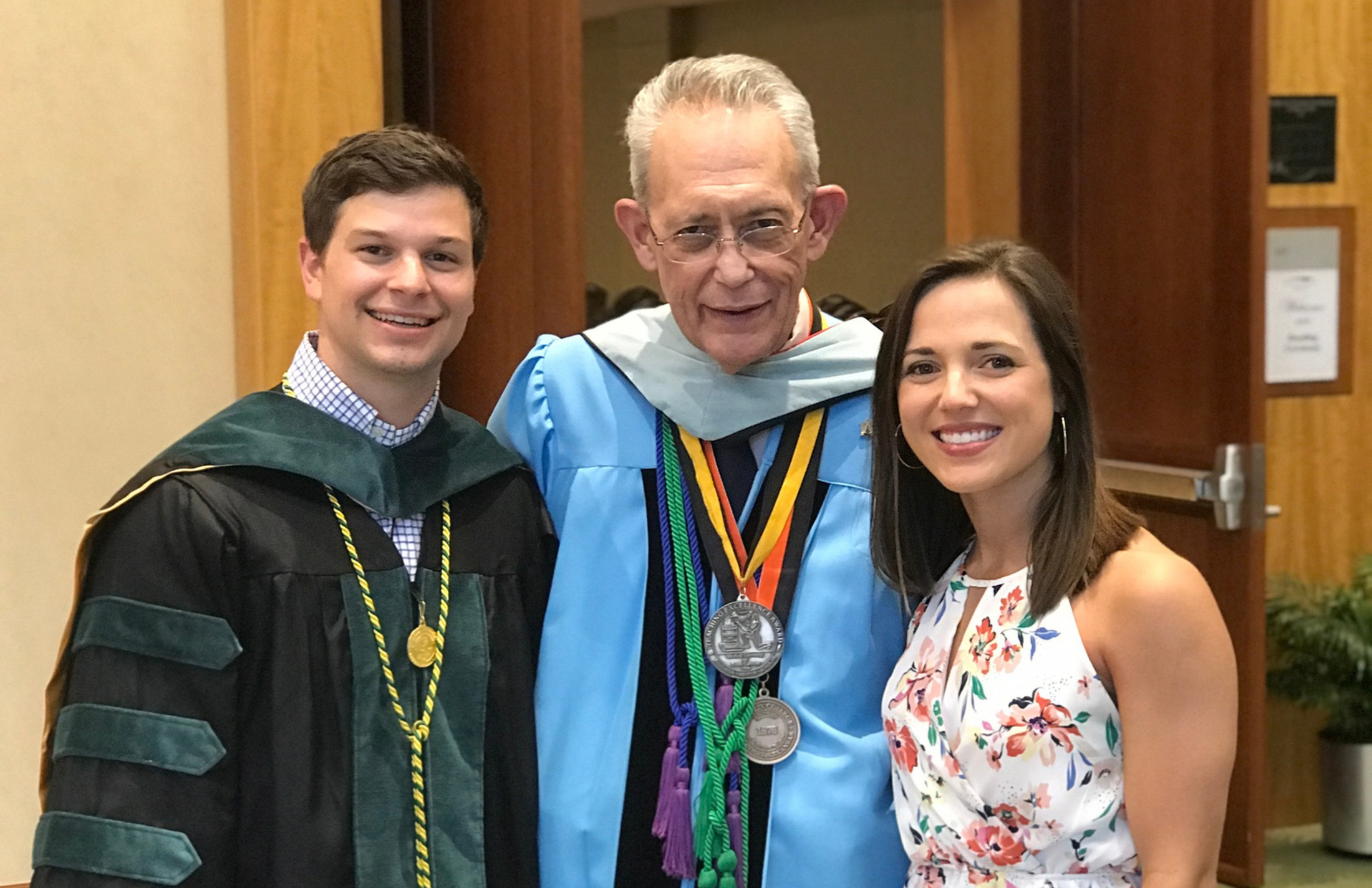 A graduate in a black gown, an older person in ceremonial robes, and a woman in a floral dress smile warmly indoors, conveying celebration and achievement.