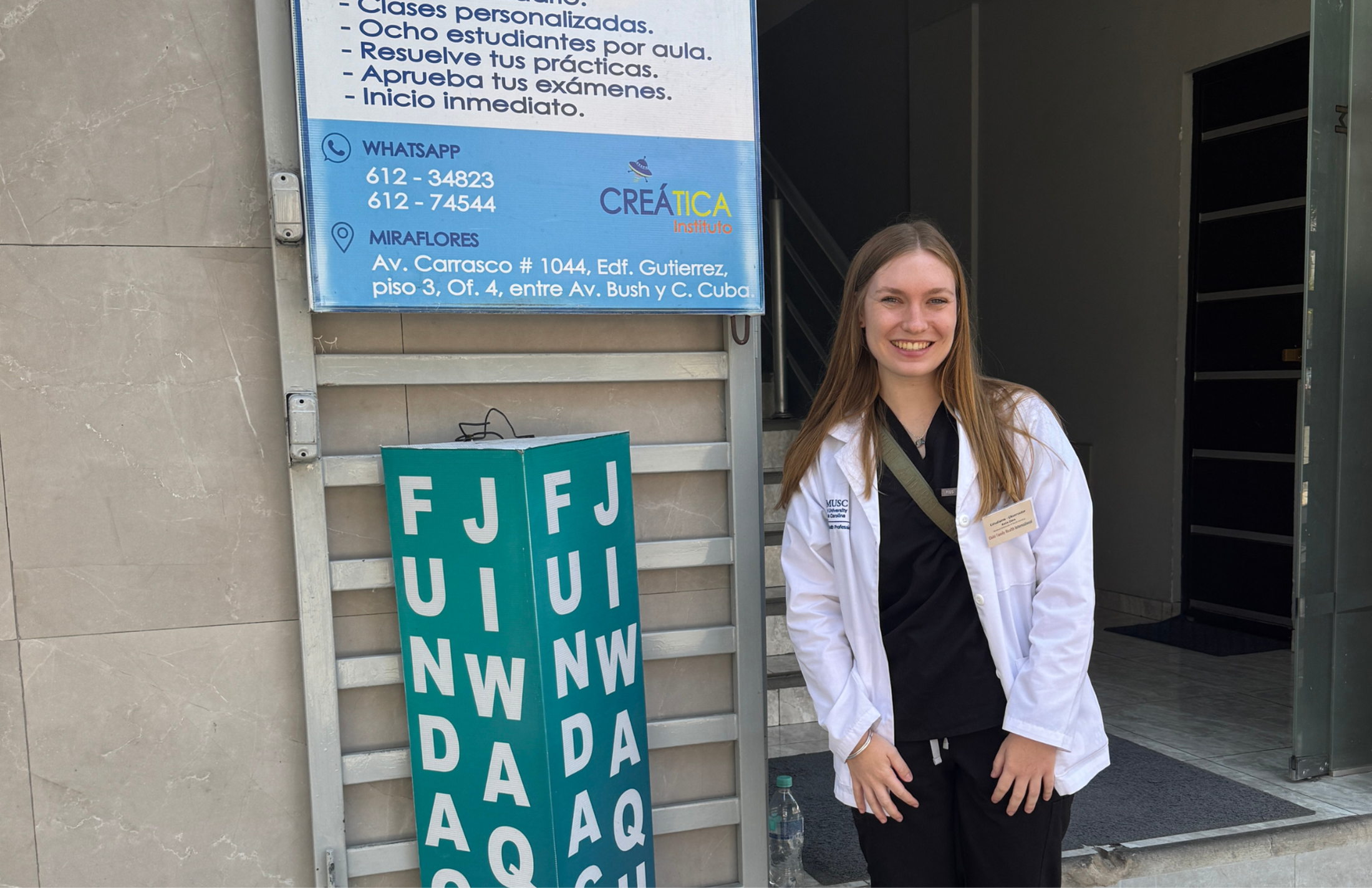 A young woman in a medical white coat stands outside a clinical site in La Paz, Bolivia, next to a sign for a local institute.