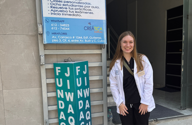 A young woman in a medical white coat stands outside a clinical site in La Paz, Bolivia, next to a sign for a local institute.