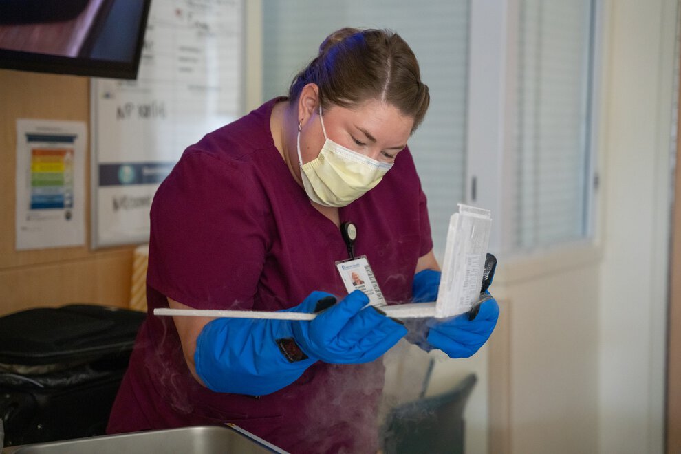 A nurse wearing a surgical mask and thick insulated gloves opens a plastic box in a hospital room.