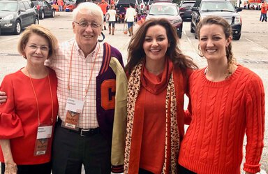 A family of four stands in a parking lot at what appears to be a tailgating event. The man is wearing a Clemson jacket. His wife and two daughters are wearing Clemson orange.
