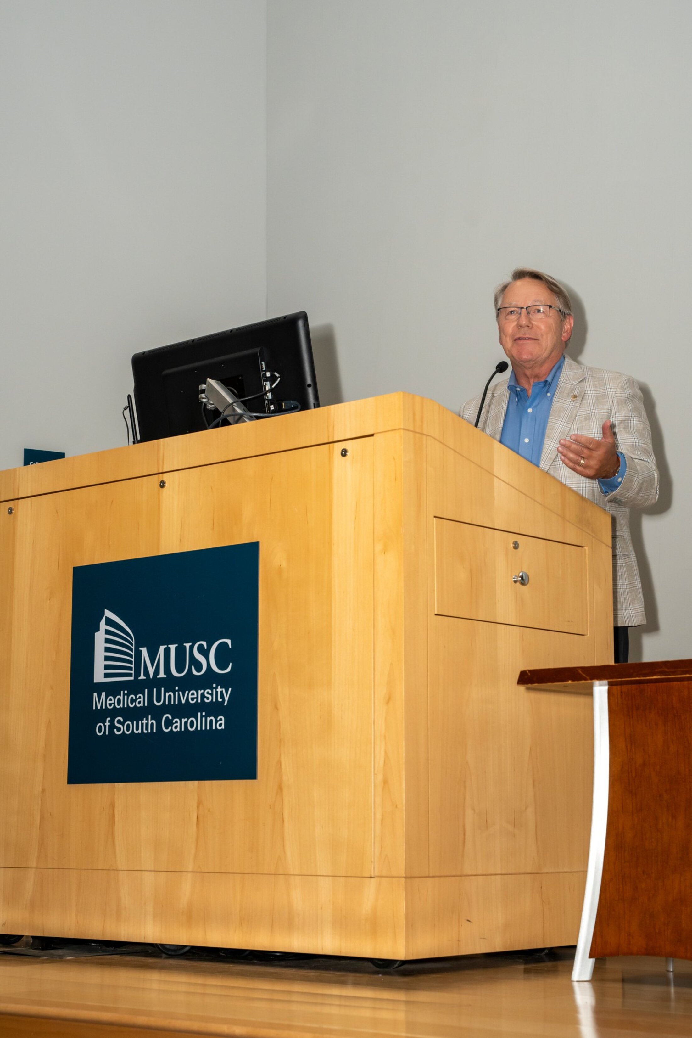 Man behind a large wooden podium wearing a blue shirt and tan jacket gestures as he speaks. The podium has the MUSC logo on it.