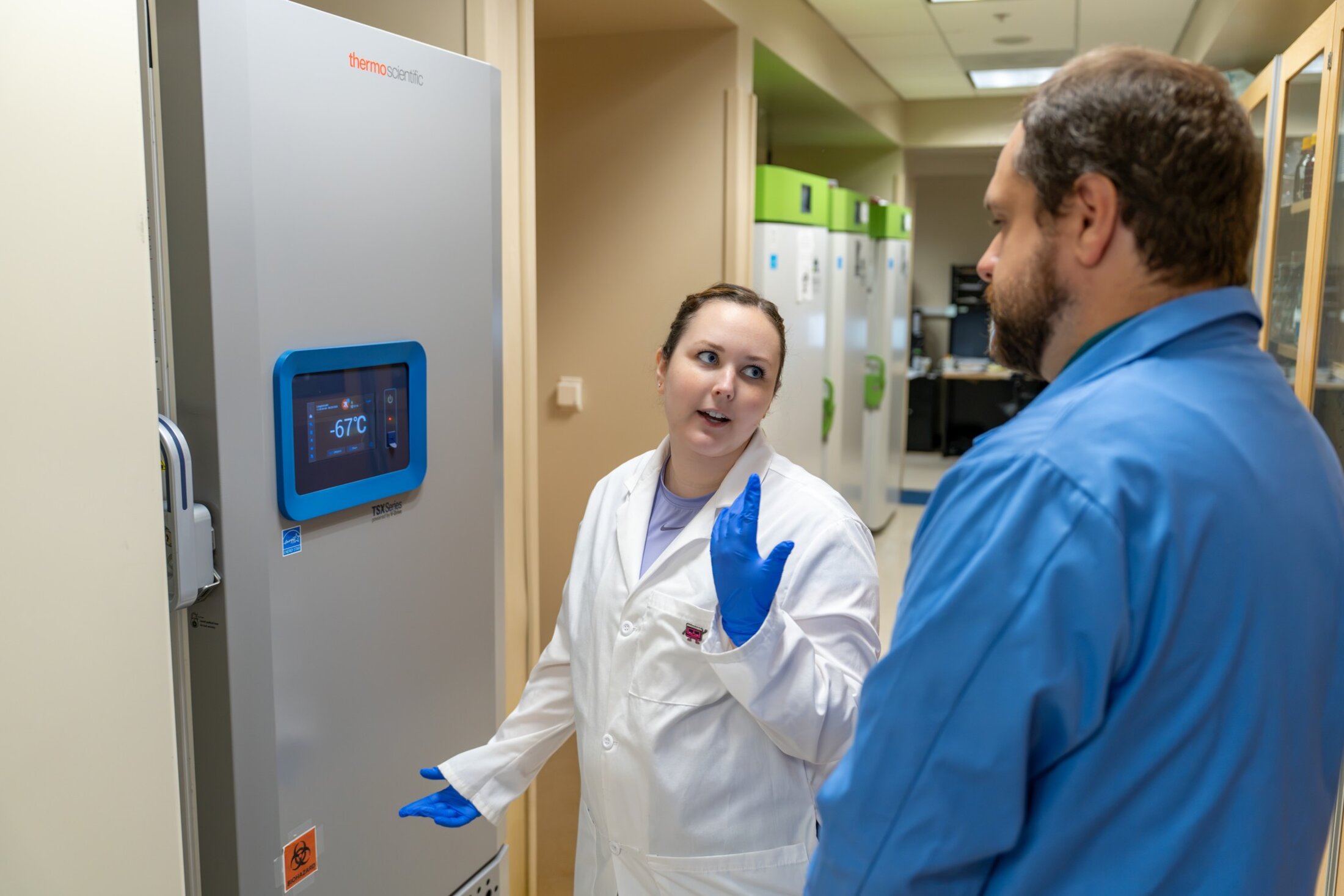 A researcher in white coat and blue gloves looks over her shoulder to chat to her mentor before opening a lab freezer that reads -67 C degrees.
